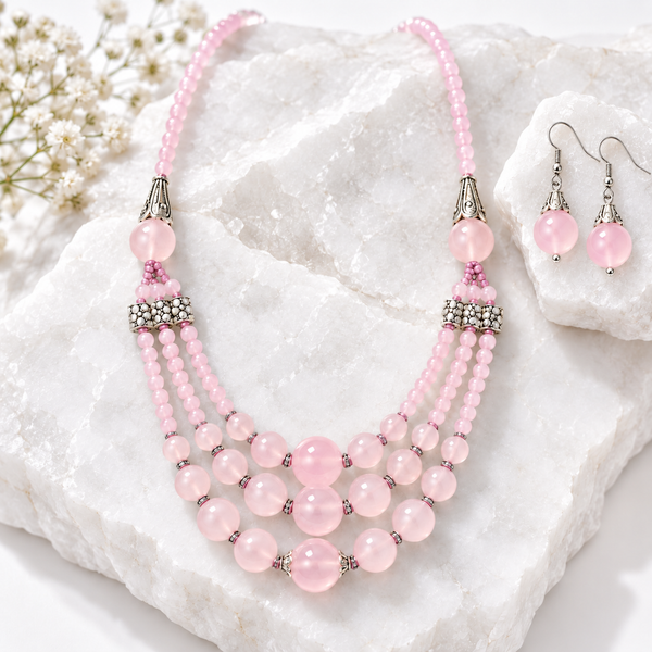 A pink rose quartz beaded statement necklace with matching pink rose quartz drop earrings, styled on white rock with small white flowers. The necklace features multiple strands of light pink rose quartz beads, silver-toned metal spacers, and crystal-like details.